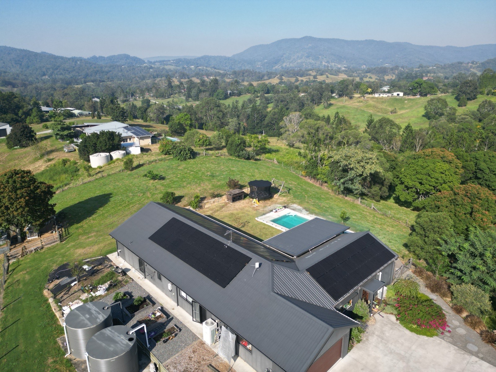 Drone view of an acreage home with rooftop solar panels, a pool and water tanks
