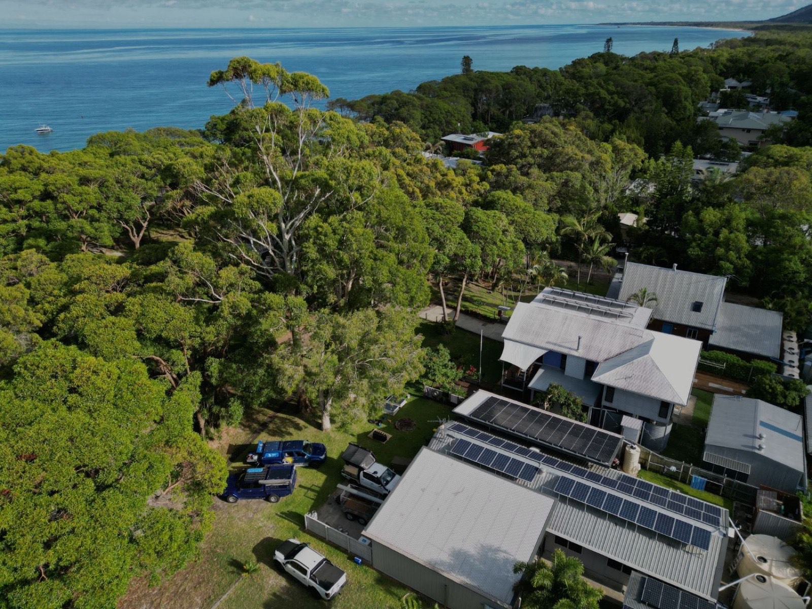 Coastal property with solar panels across several roofs and water tanks, surrounded by bush